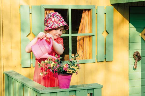 Mädchen gießt Blumen vor buntem Holzspielhaus